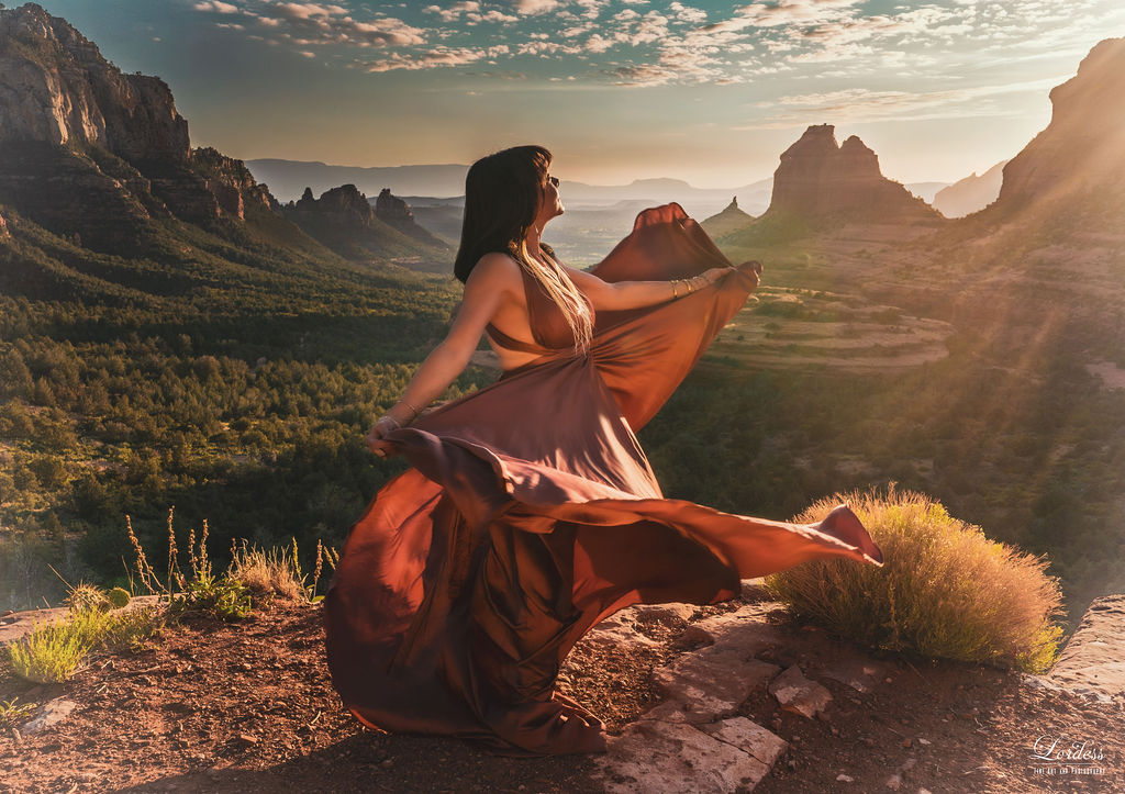 Terii in a flowing pink and rust silk dress on a Sedona cliff at golden hour, dress catching the wind, red rock vistas behind her